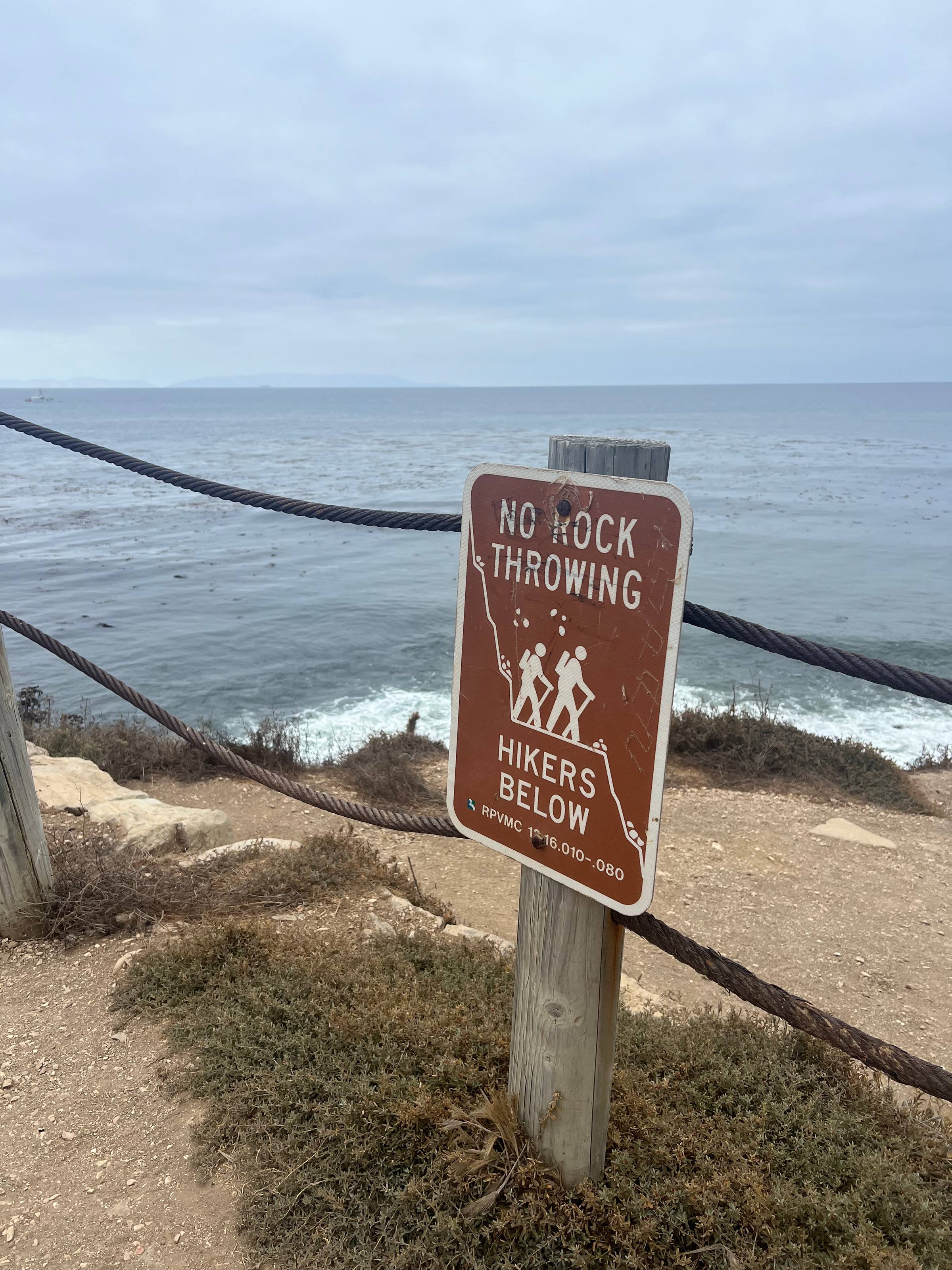 brown sign on post reading "no rock throwing hikers below" on an ocean cliff