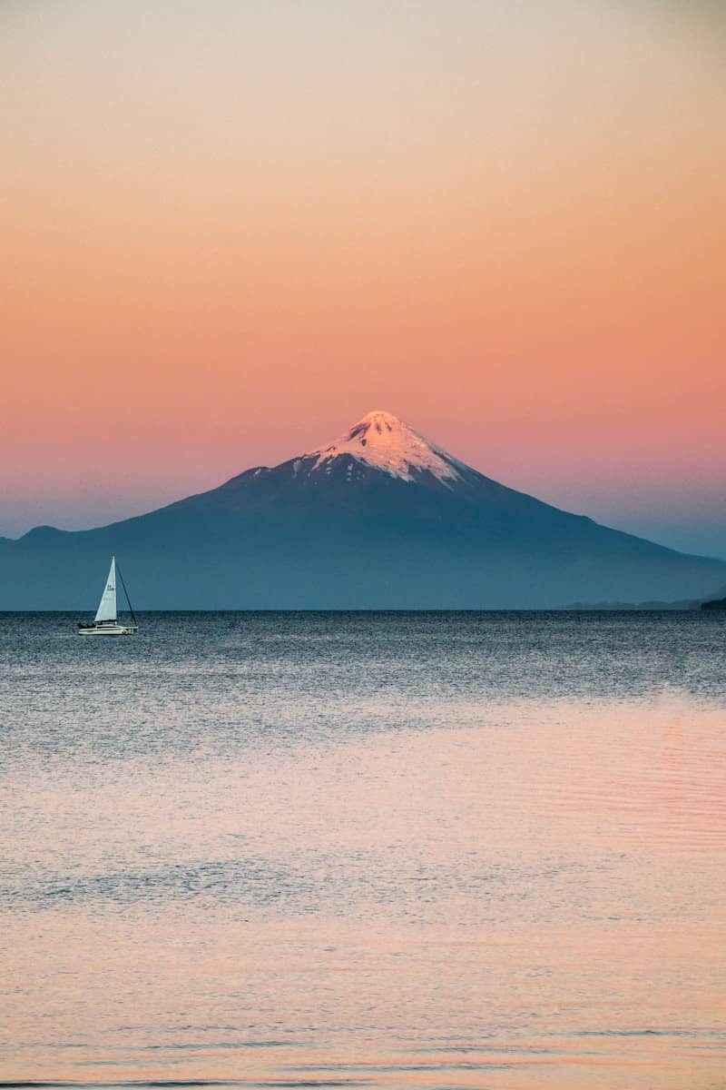 Sailboat on calm water with snow-capped mountain at sunset.
