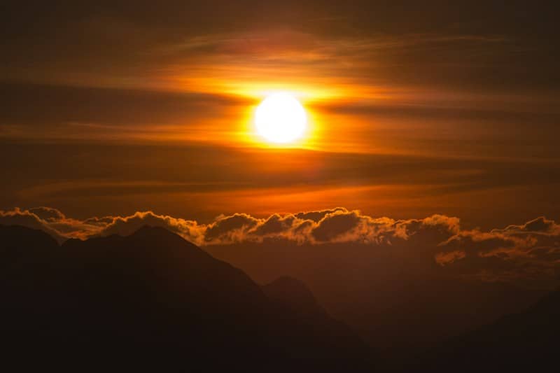 A bright low sun above dark mountain silhouettes and clouds in an orange sky
