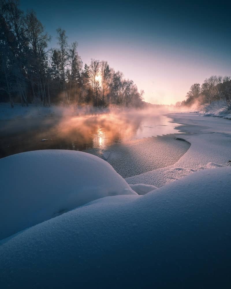 Misty river flows through a snowy forest at sunrise.