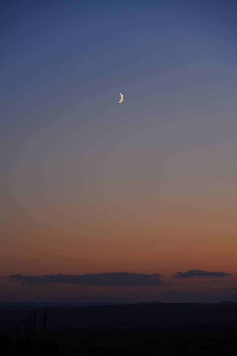 A crescent moon hangs above a colorful sunset.