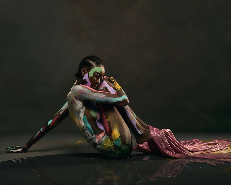 A person with colorful body paint curled on a dark reflective surface