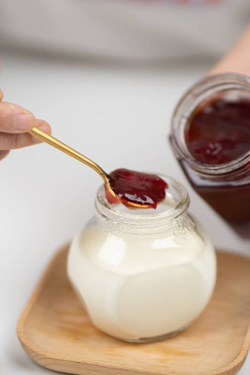 Creamy homemade yogurt in a small mason jar topped with jam being spooned over the top