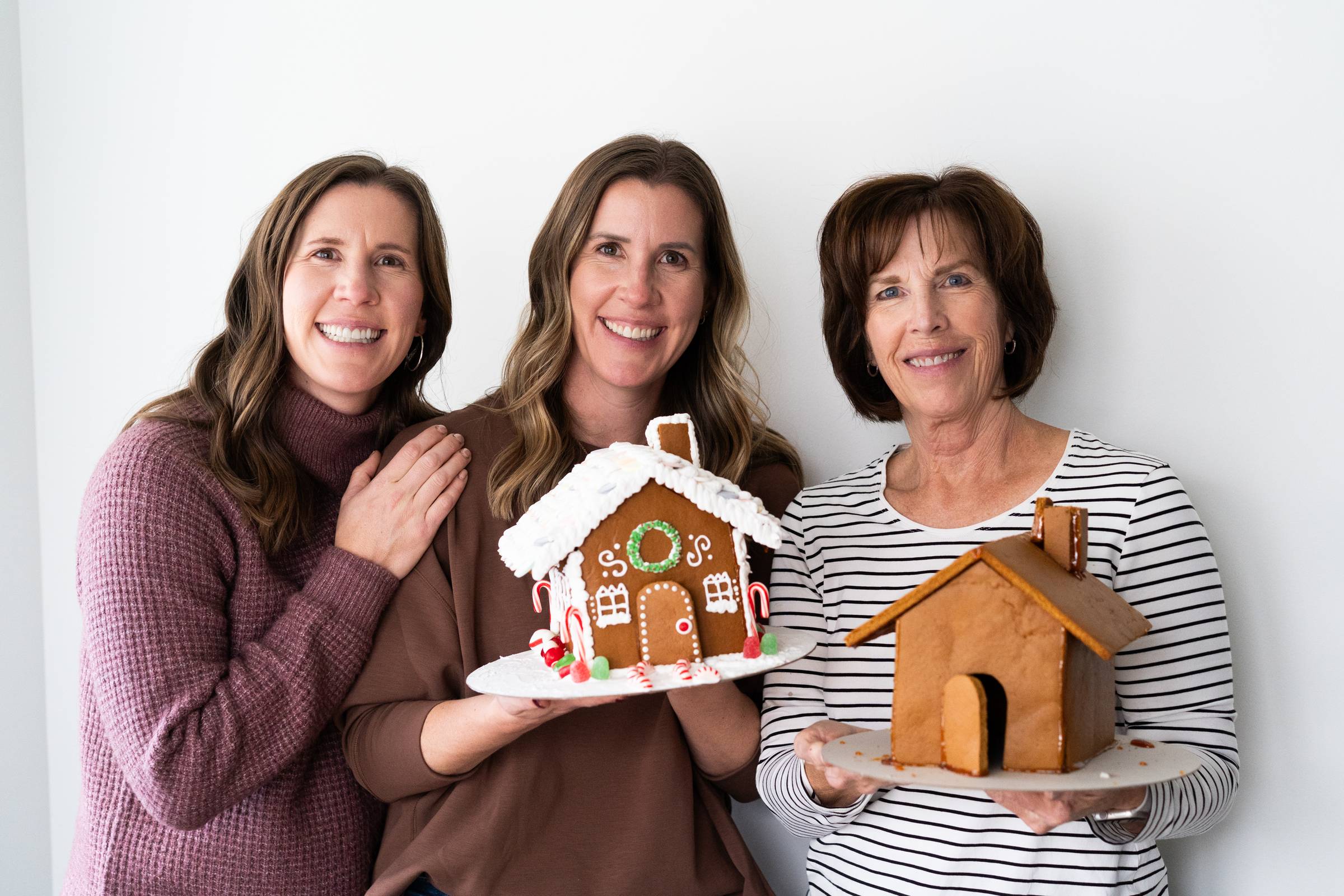 Liz, Lauren, and their Mom posing with a finished gingerbread house and a non decorated gingerbread house. 