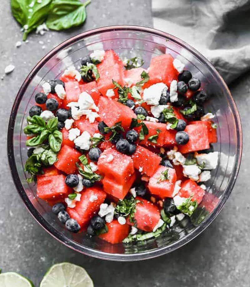 Watermelon Salad served in a clear bowl with herbs in the background.