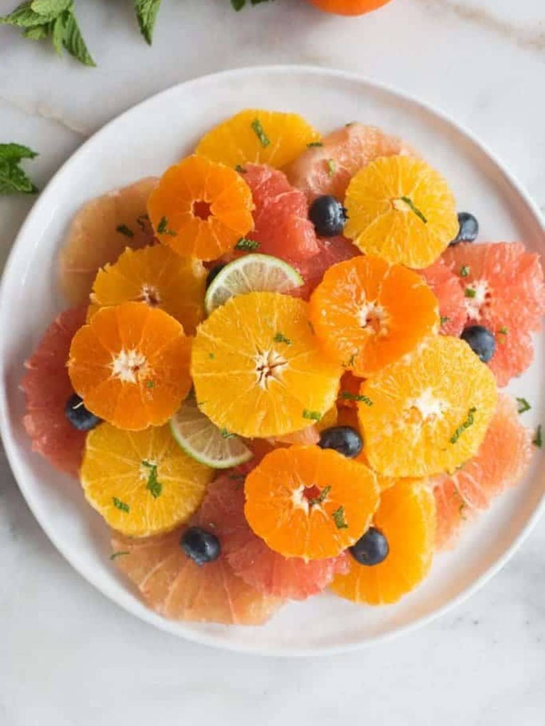 sliced oranges, grapefruit, and other citrus stacked on a white plate and garnished with mint and blueberries. 