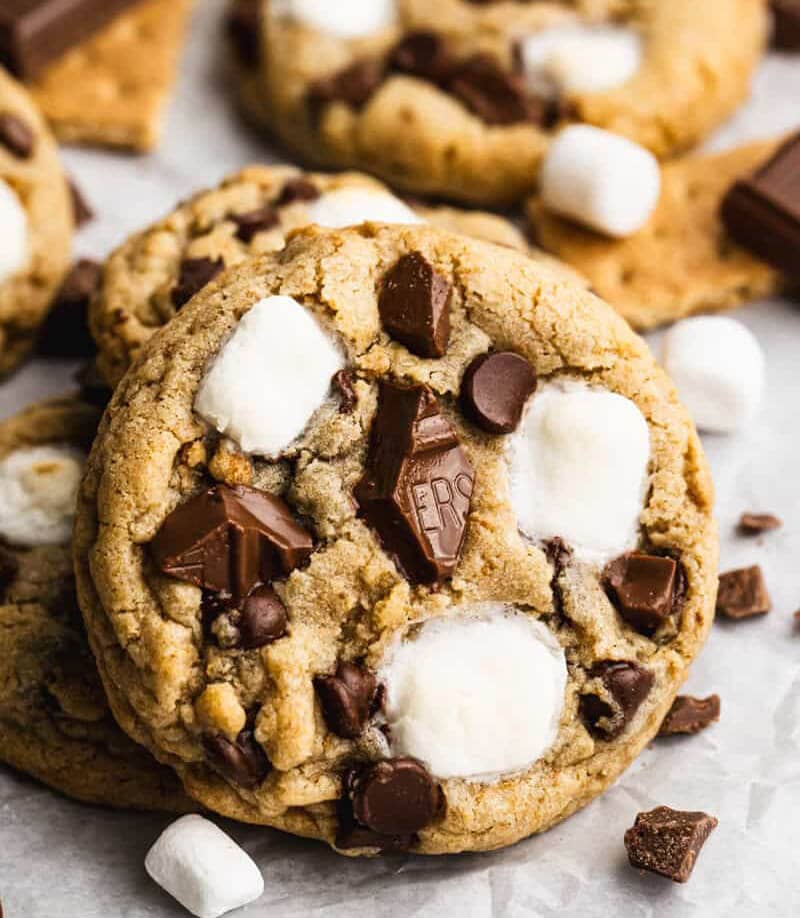S'mores Cookies with one in focus and a random pile of more cookies in the background. Mini marshmallows and bits of chocolate are scattered around. 