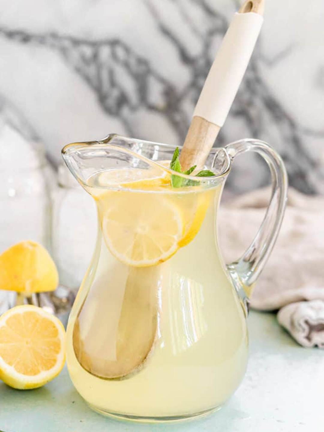 Lemonade in a clear glass pitcher garnished with sliced lemons and a mint leaf.