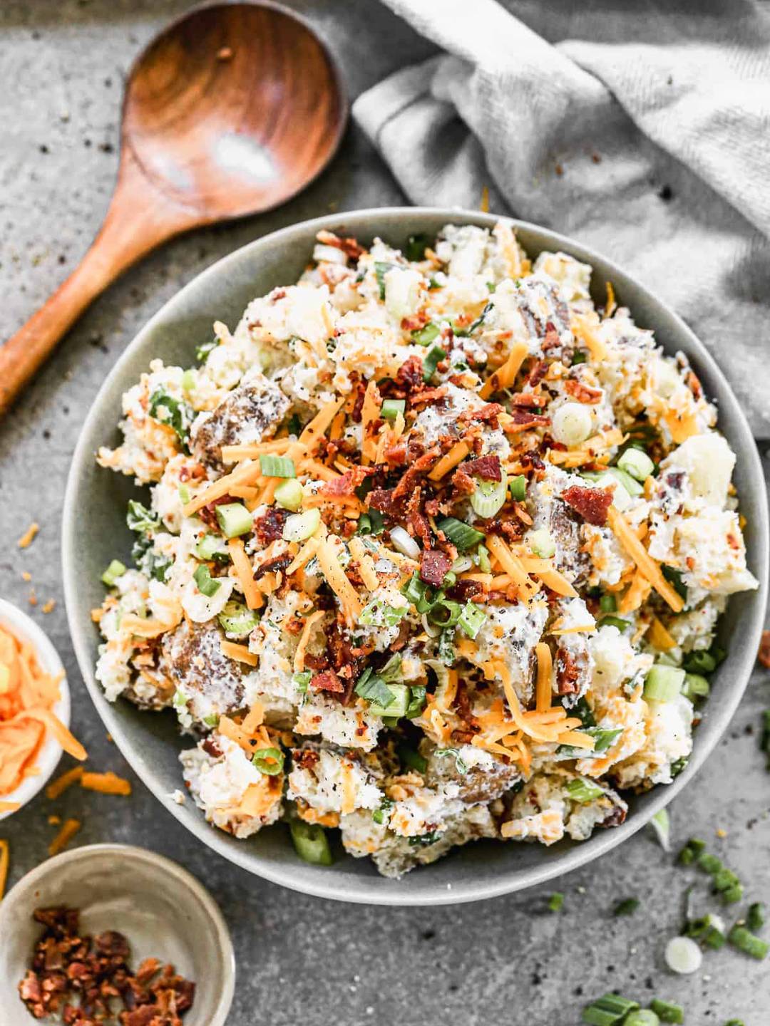 Baked Potato Salad in a grey bowl with ingredients and a wooden spoon surrounding.