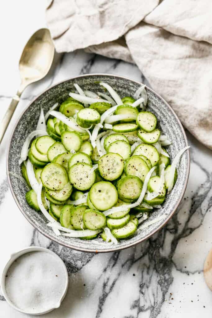 Cucumber Onion Salad served in a grey bowl and surrounded by a cream colored kitchen towel and a gold serving spoon.
