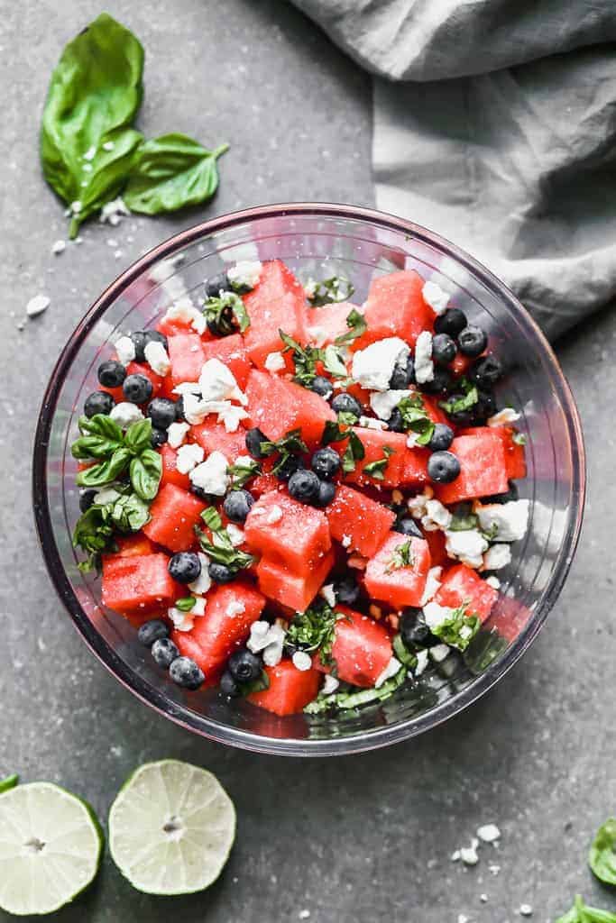 Watermelon Salad includes feta cheese, blueberries, and fresh basil, with a hint of lime. Served in a clear bowl.