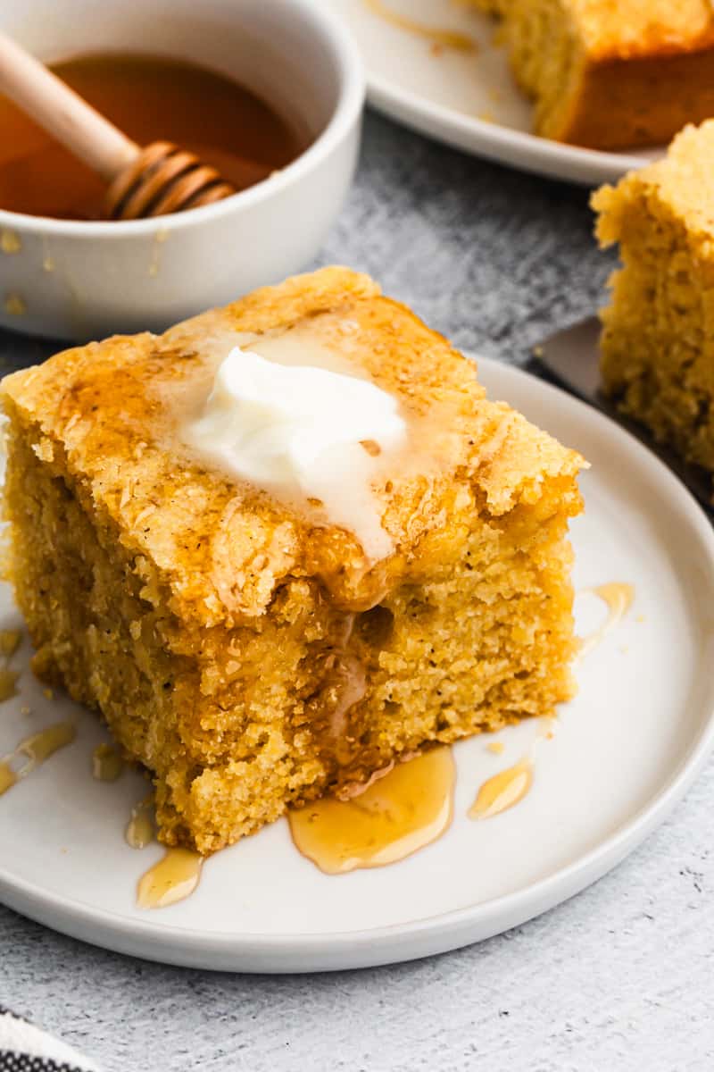 Buttermilk Cornbread served on a white plate drizzled with honey and butter with honey and more servings in the background. 