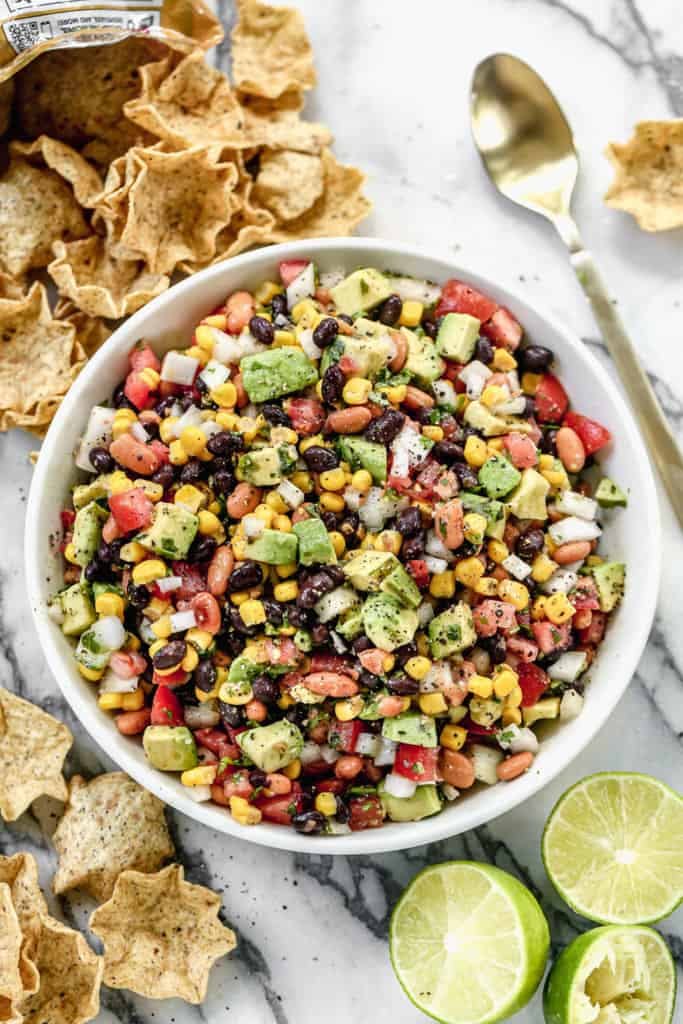 Cowboy Caviar is a mix of beans, onion, cilantro, avocado, and fresh veggies served in a white bowl and tortilla chips