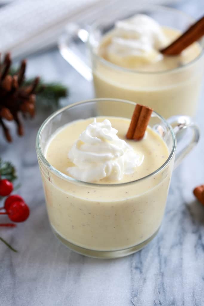 two cups of eggnog in clear mugs topped with whipped cream and a cinnamon stick