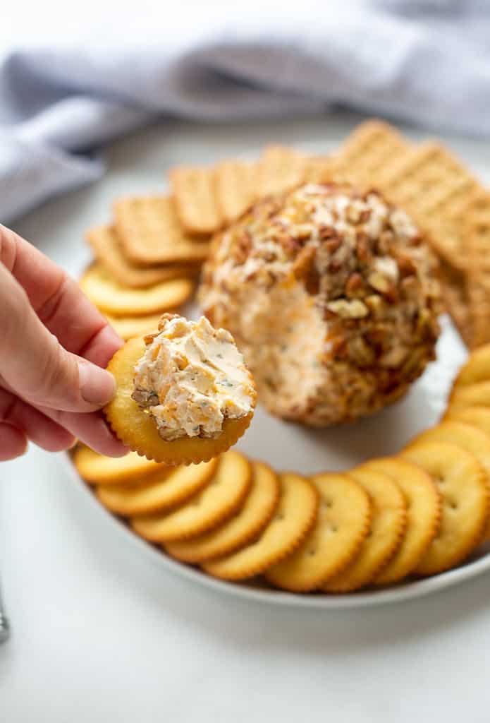 Classic Cheese Ball arranged on a white tray and pictured with a hand holding a cracker with the spread on it.