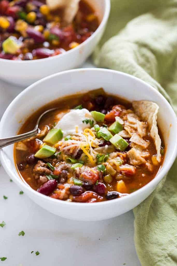 Taco Soup in a white bowl topped with sour cream, avocado, and shredded cheddar cheese. Serve with tortilla chips