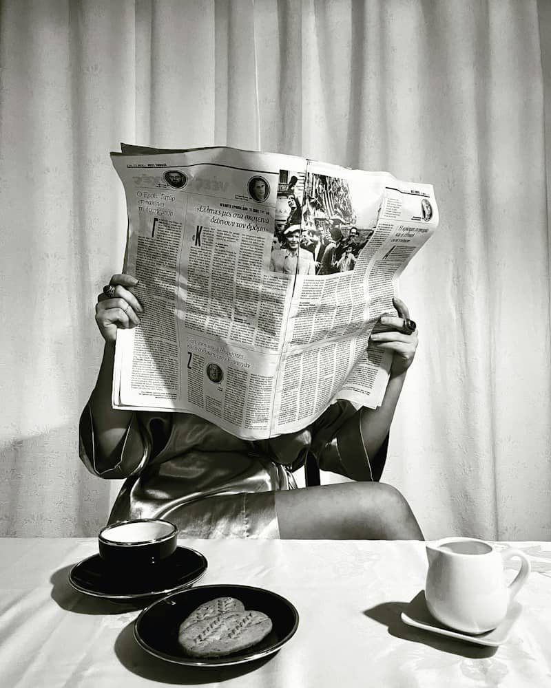 A person in a silk robe reading a newspaper at a table with coffee
