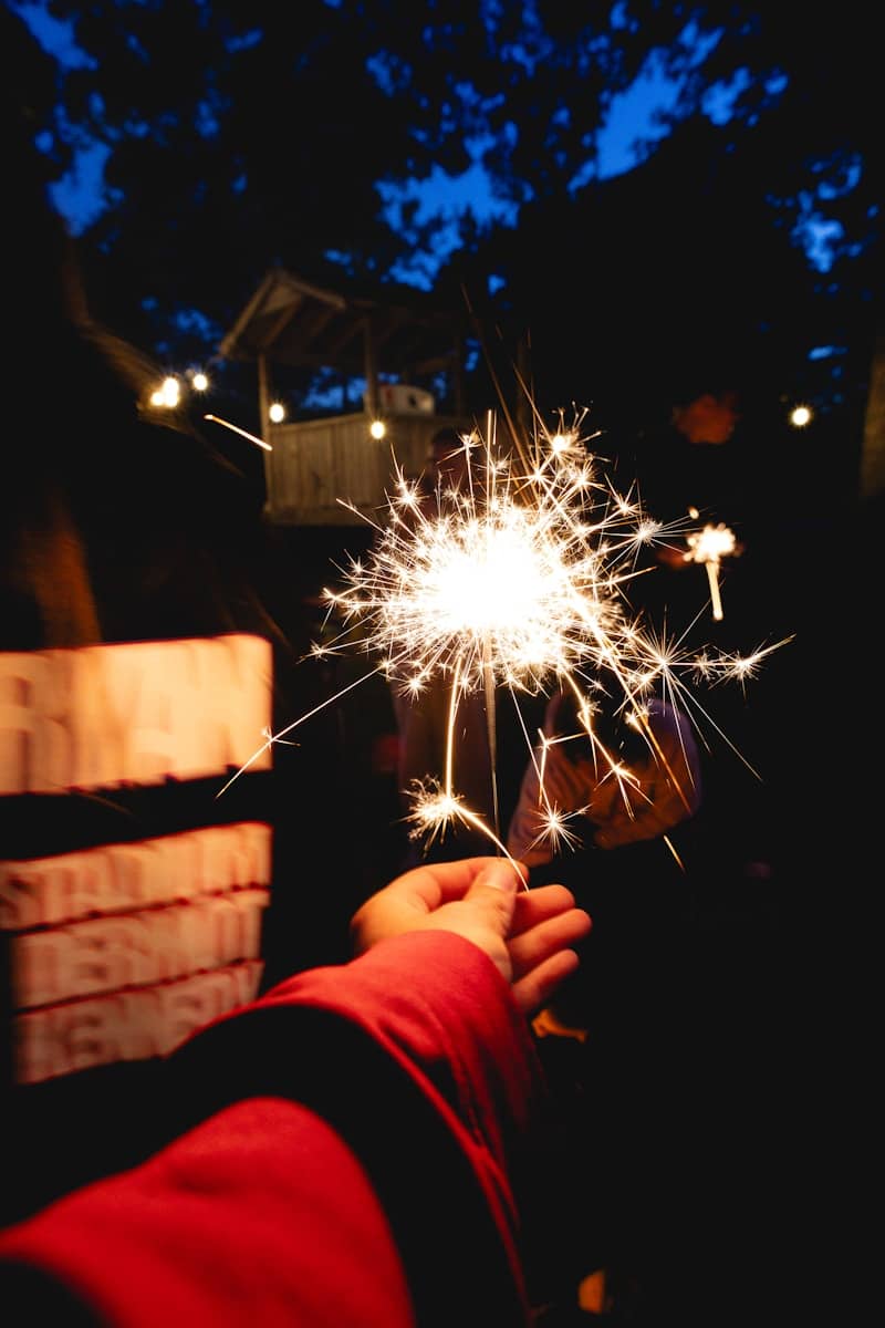 A hand holds a sparkling lit sparkler at night.