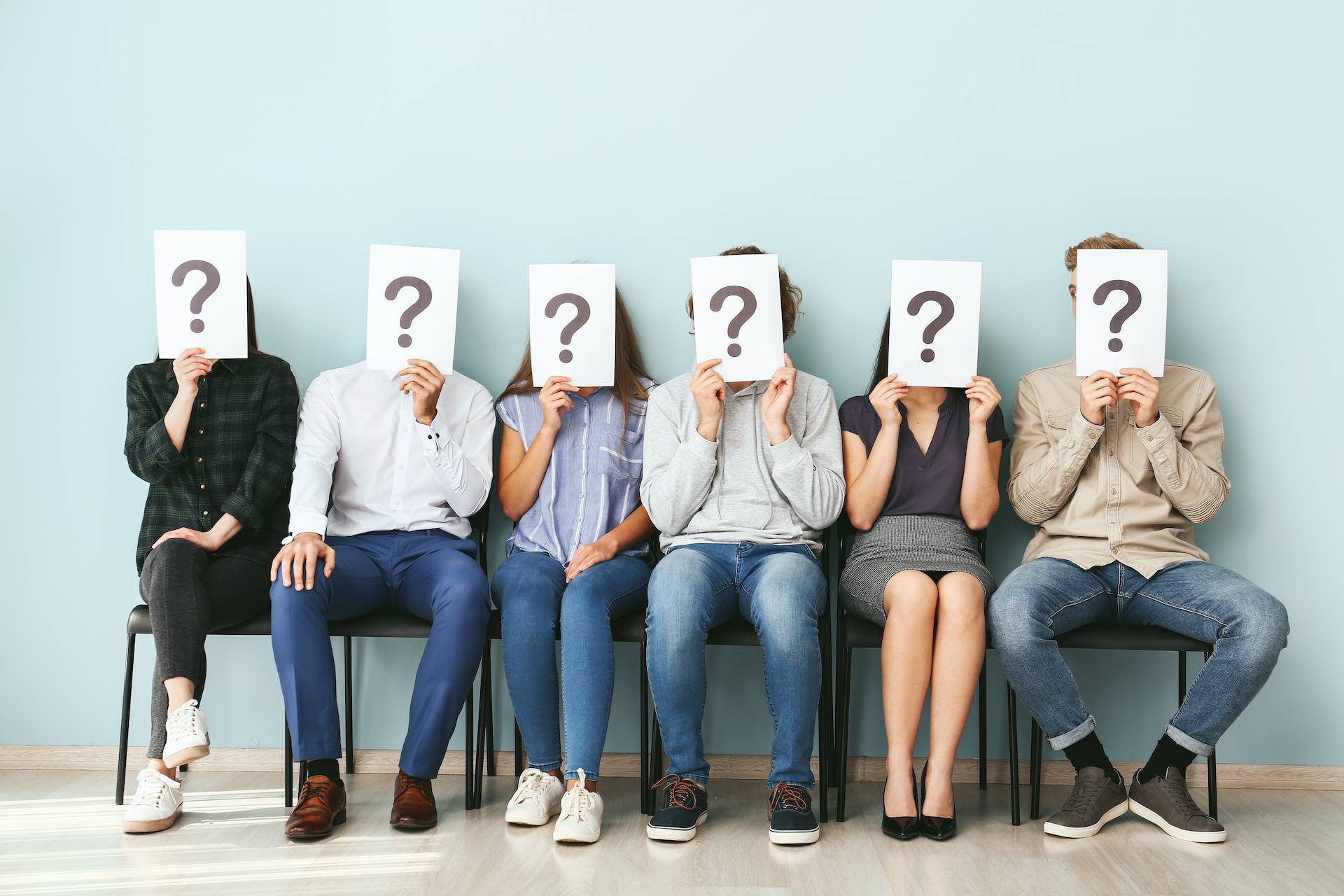 6 people sitting in chairs in a line, each holding a piece of paper in front of their face with a question mark on it.