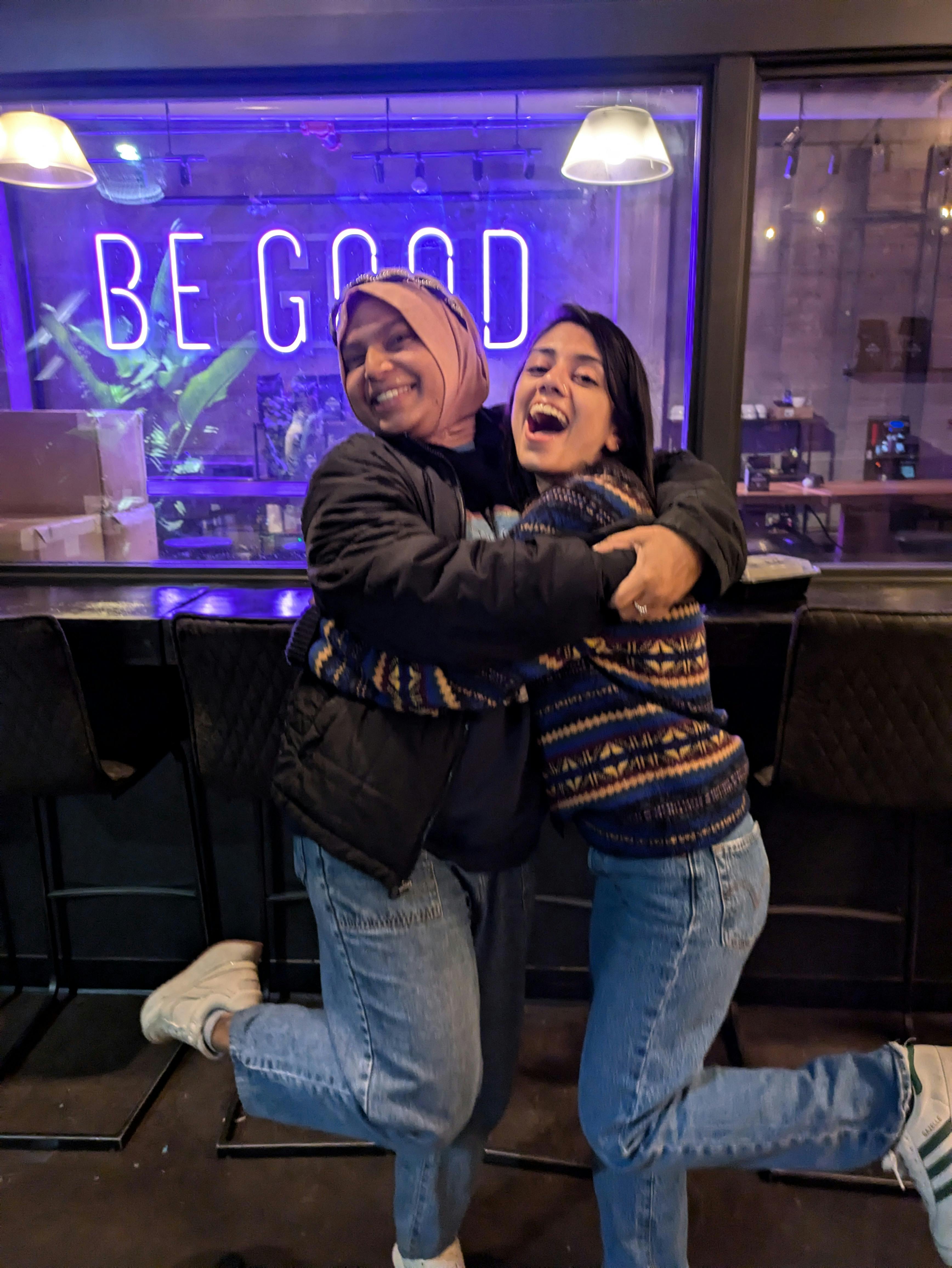 2 soapbox members Daaniya and Medha hugging it out in front of a neon sign that says BE GOOD