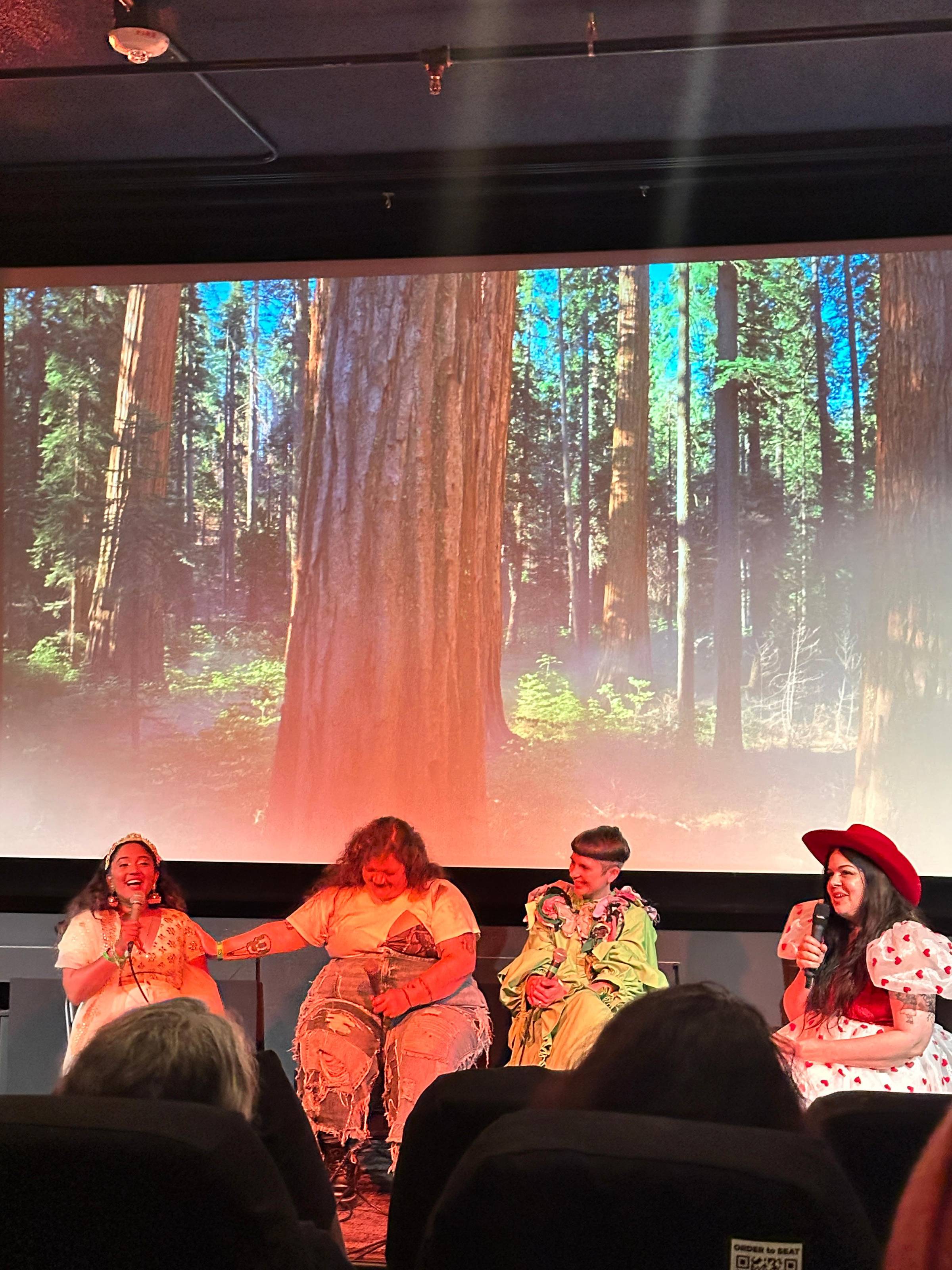 us 4 podcast people laughing. idk at what. behind us is a projectorscreen of a redwood forest