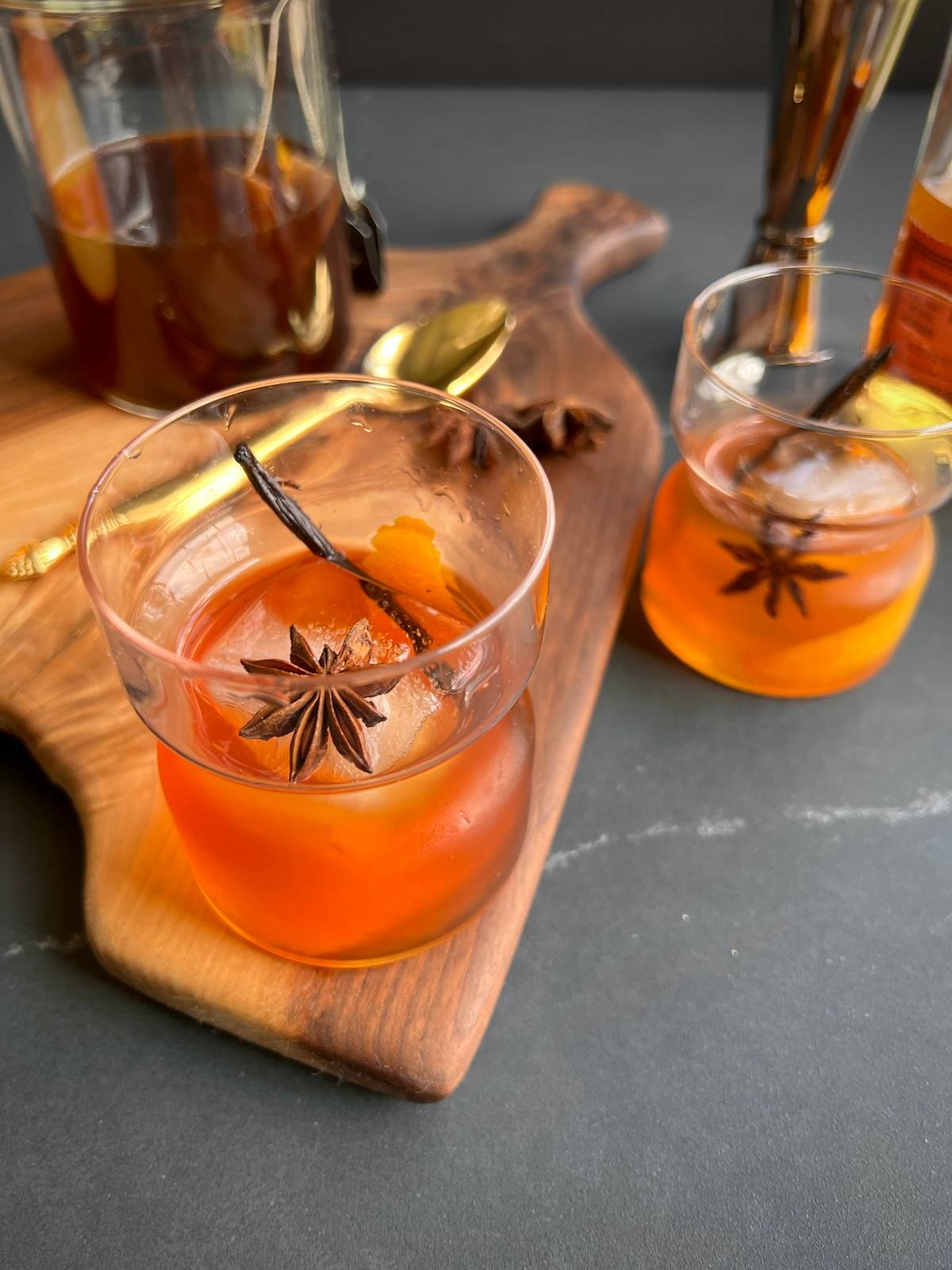 two glasses of vanilla chai old fashioned are sitting on a rustic wooden board