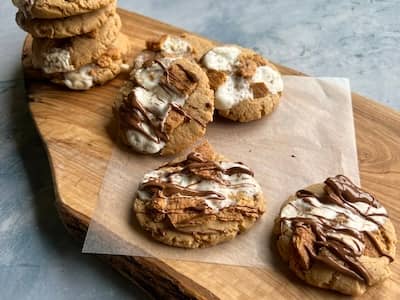 peanut butter s'mores cookies on a wooden serving board. one cookie is front and center to show off its chocolate drizzle and there is a tall stack of cookies in the background.