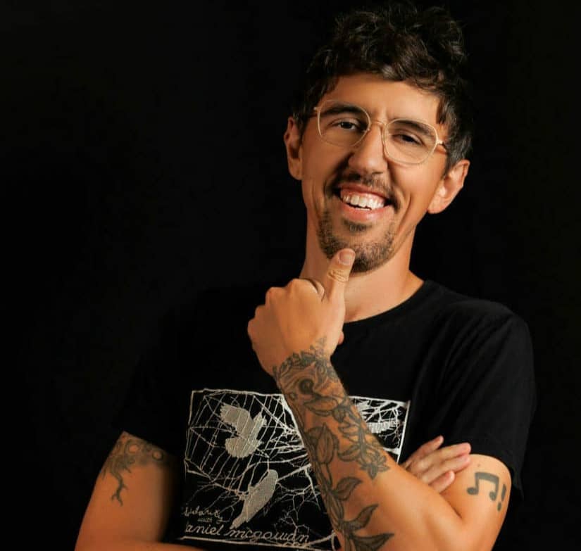 A person with short, dark hair and a goatee is smiling and looking at the camera. They are wearing glasses and a black t-shirt with a white graphic design. Their arms are crossed, and they have tattoos on both arms. One hand is touching their chin. The background is dark.