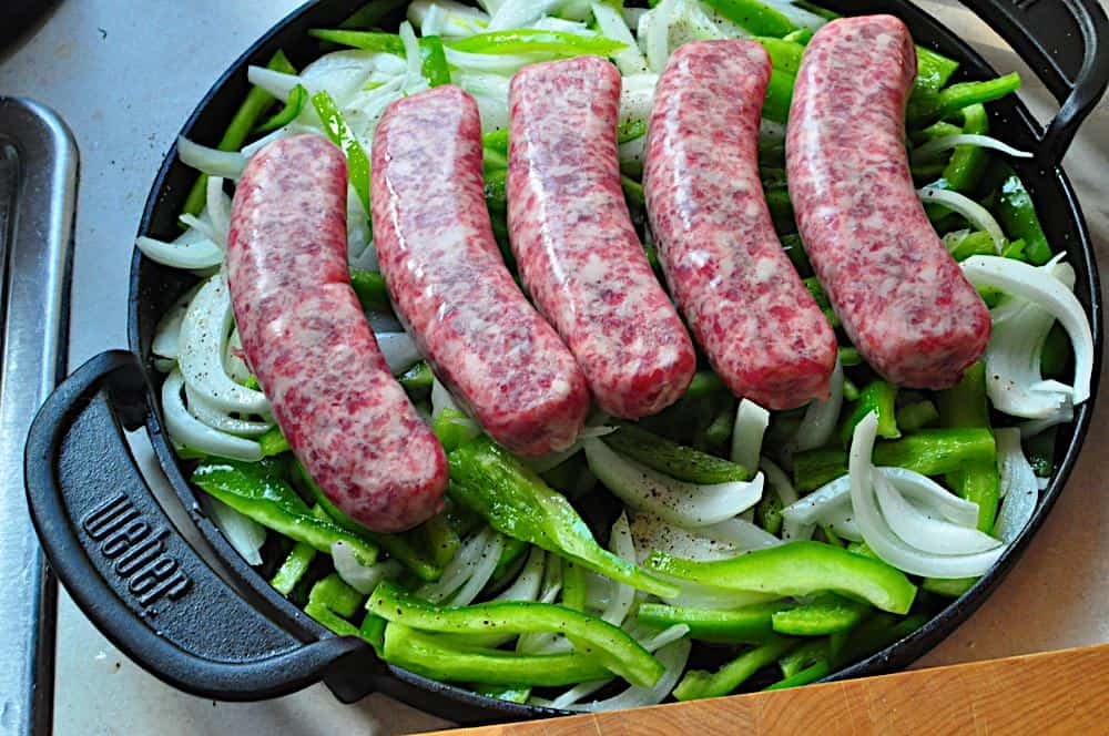 A pan of uncooked brats on top of peppers and onions