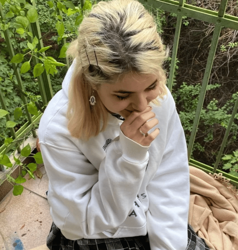 A girl with bleached blonde hair sitting on her knees on a balcony with green railing. She is wearing a white hoodie and behind her it's lush green with trees and plants. 