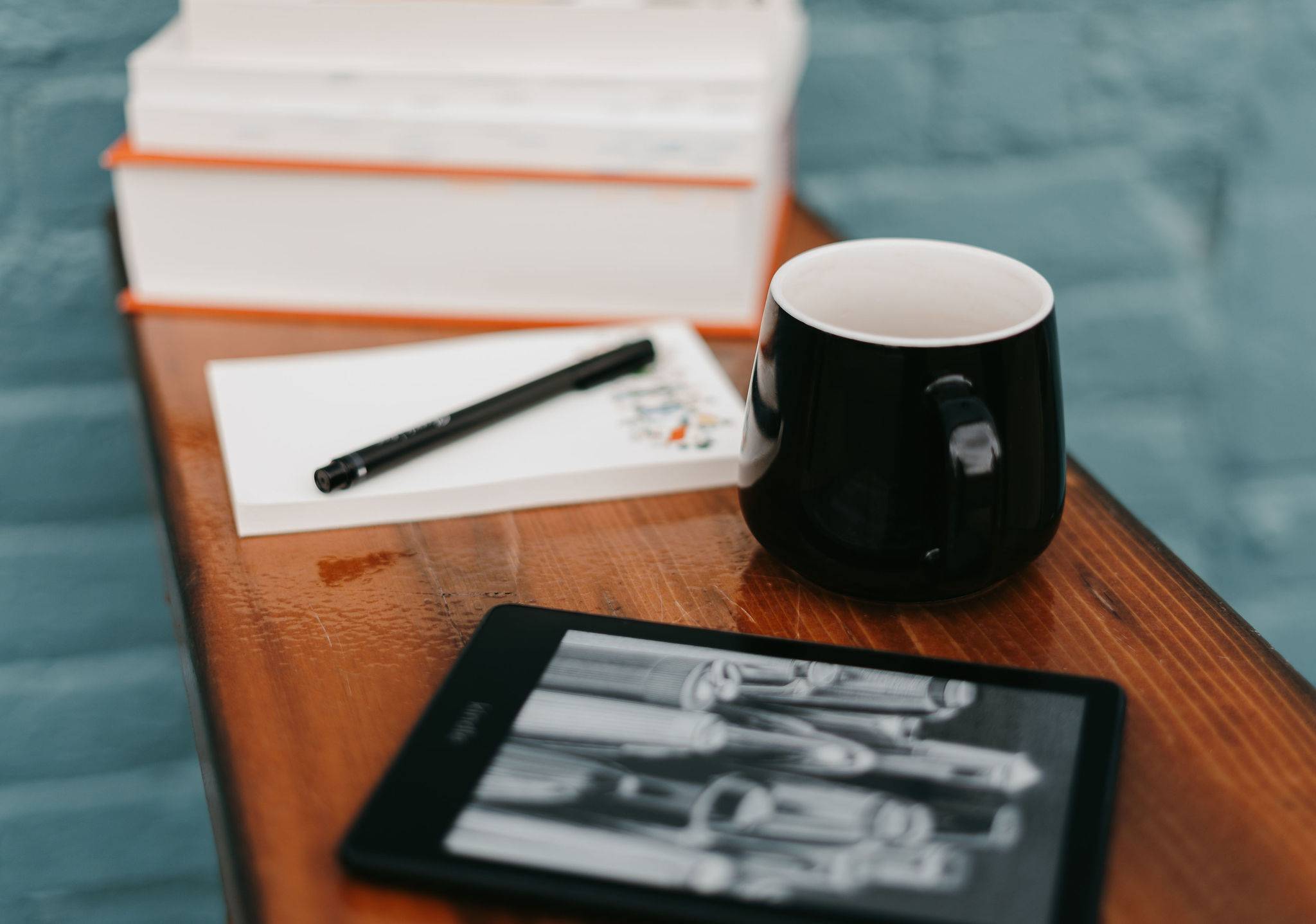 A stack of books, a notepad, an e-reader, and a coffee cup sit on a wooden bench.