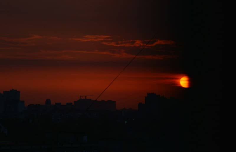 A glowing orange sun partially obscured by clouds above a dark city skyline