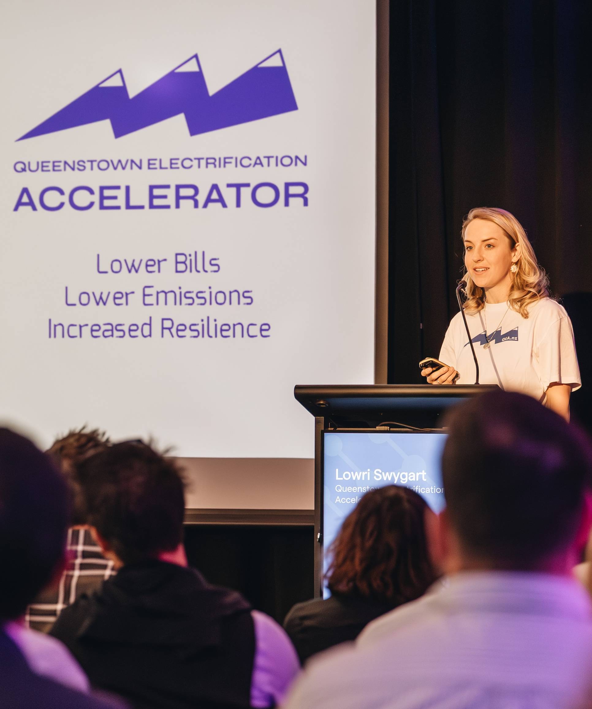 Young woman speaking at lectern with large projector slides behind reading "Queenstown Electrification Accelerator: Lower bills, lower emissions, increased resilience"