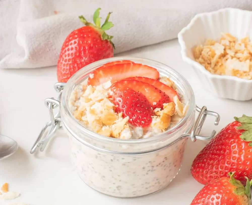 Strawberry cheesecake overnight oats in a glass jar topped with sliced strawberries and graham cracker crumbs.