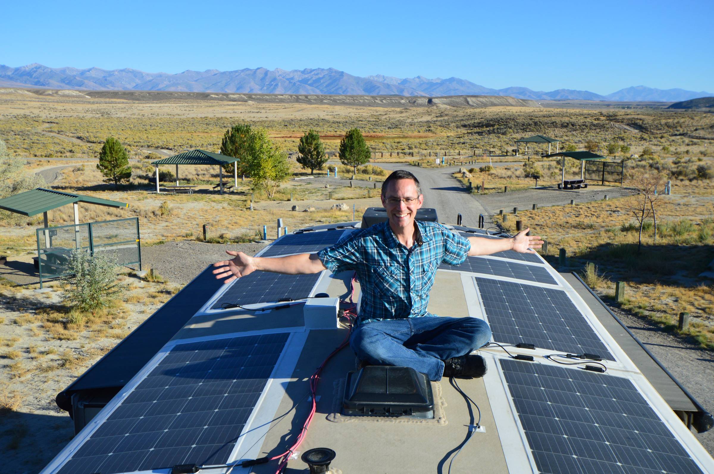 A full-time RVer sits on the roof of a fifth wheel RV surrounded by rooftop solar panels, with arms outstretched and mountain views in the distance. This image showcases an RV solar system that helps power off-grid camping and boondocking, making it easie