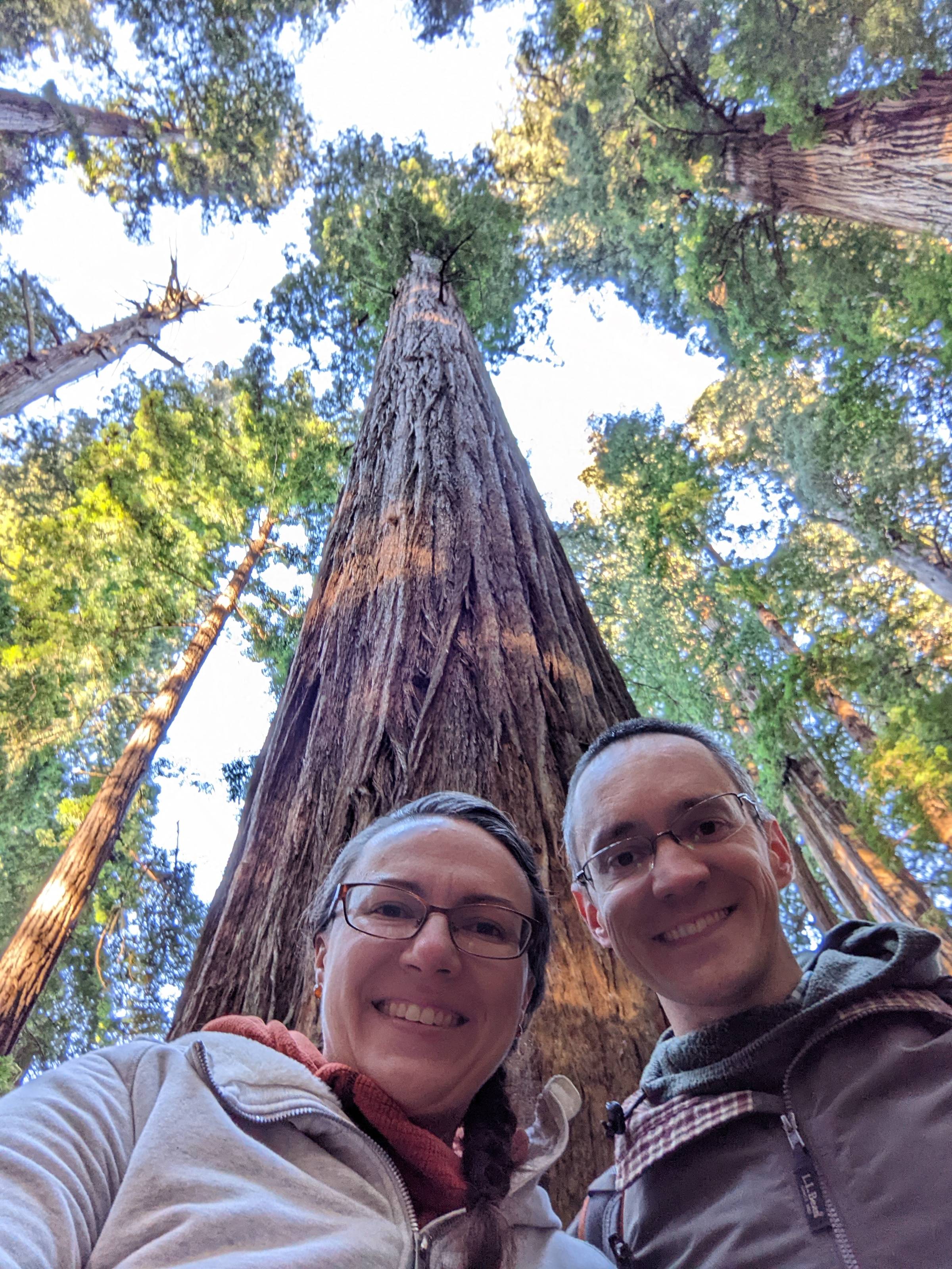 A couple smiles for a selfie beneath a towering coast redwood tree in Redwood National Park, with the massive trunk rising high above and a canopy of ancient trees filling the sky. The perspective highlights the incredible size of the old-growth redwoods,
