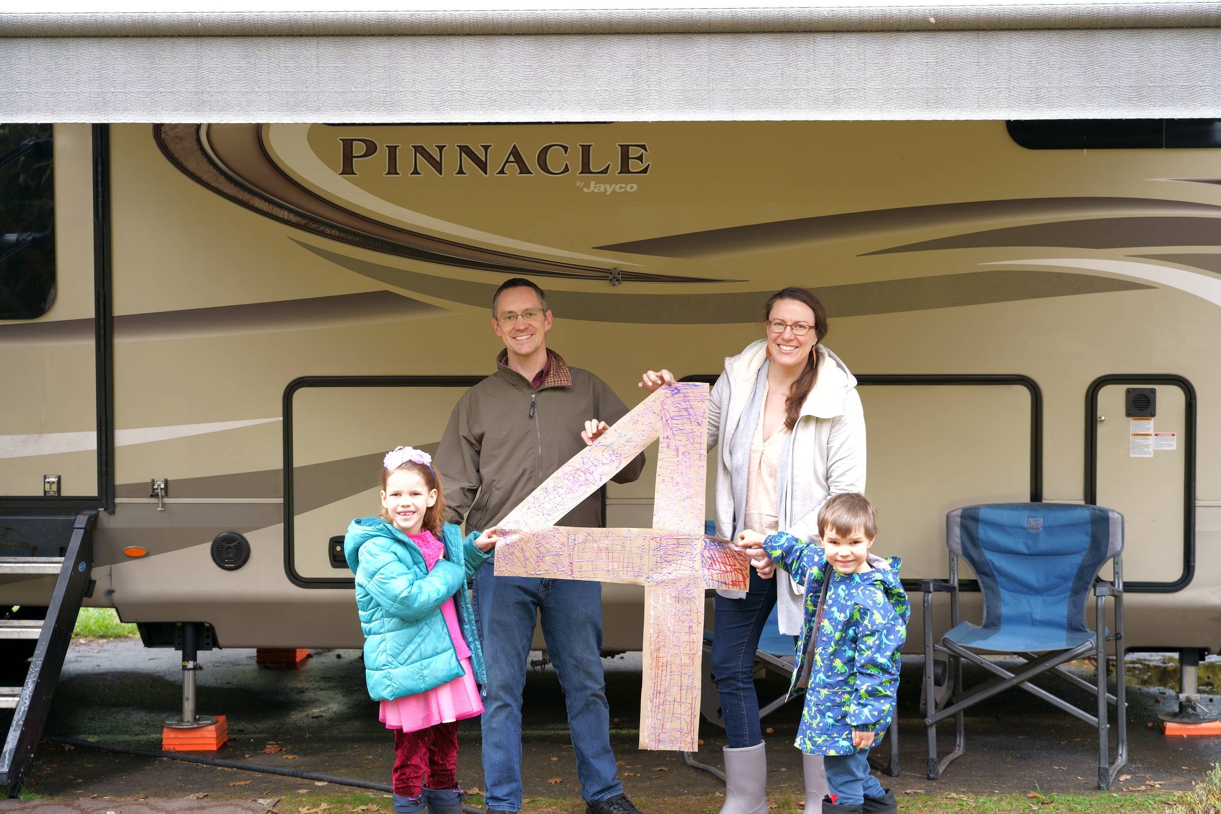 A family of four stands in front of their fifth-wheel RV holding a large handmade number four to celebrate four years of full-time RV living. The parents and children smile together at their campsite, marking an important milestone in their RV travel jour