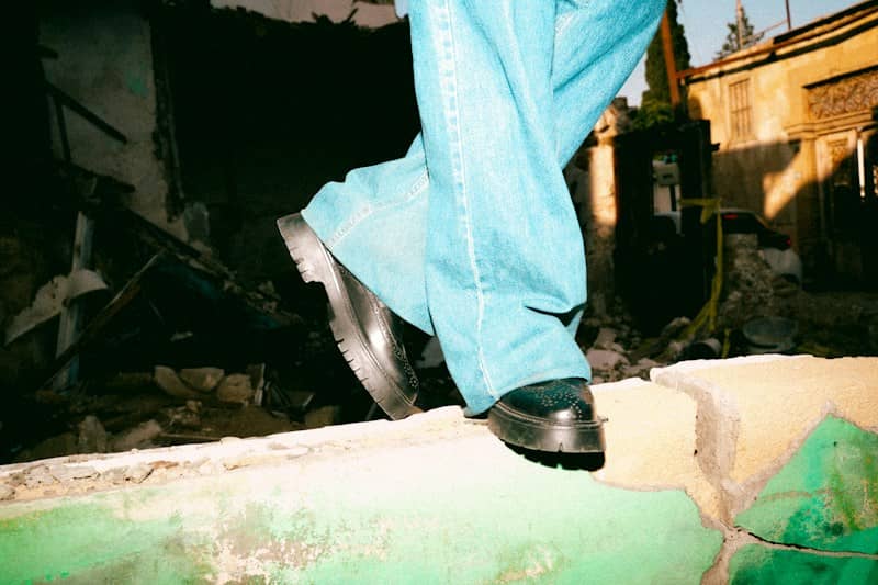 A person in wide-leg blue jeans and black brogues standing on a low green wall