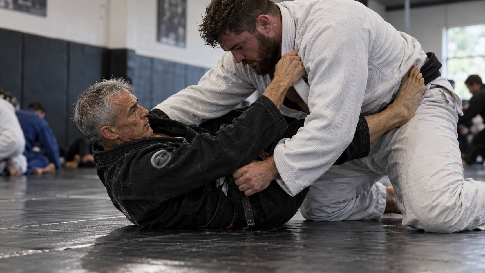A smaller older jiu-jitsu practitioner using guard and frames against a larger opponent on the mat.