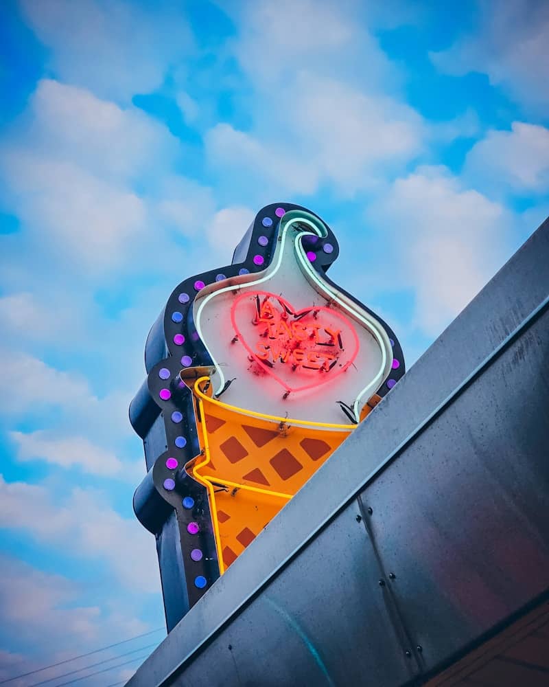 A neon ice cream cone sign with a heart against a blue cloudy sky