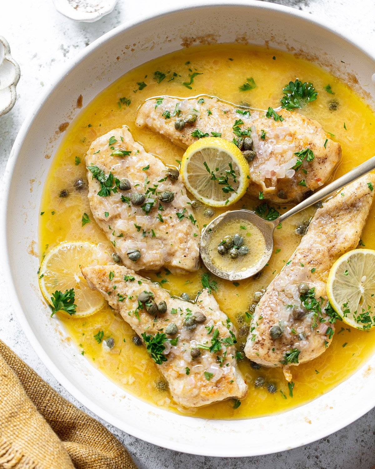 Chicken breasts in a chicken piccata are garnished with capers, parsley, and lemon slices. The dish is served in a white pan on a light surface, with a spoon resting in the sauce.