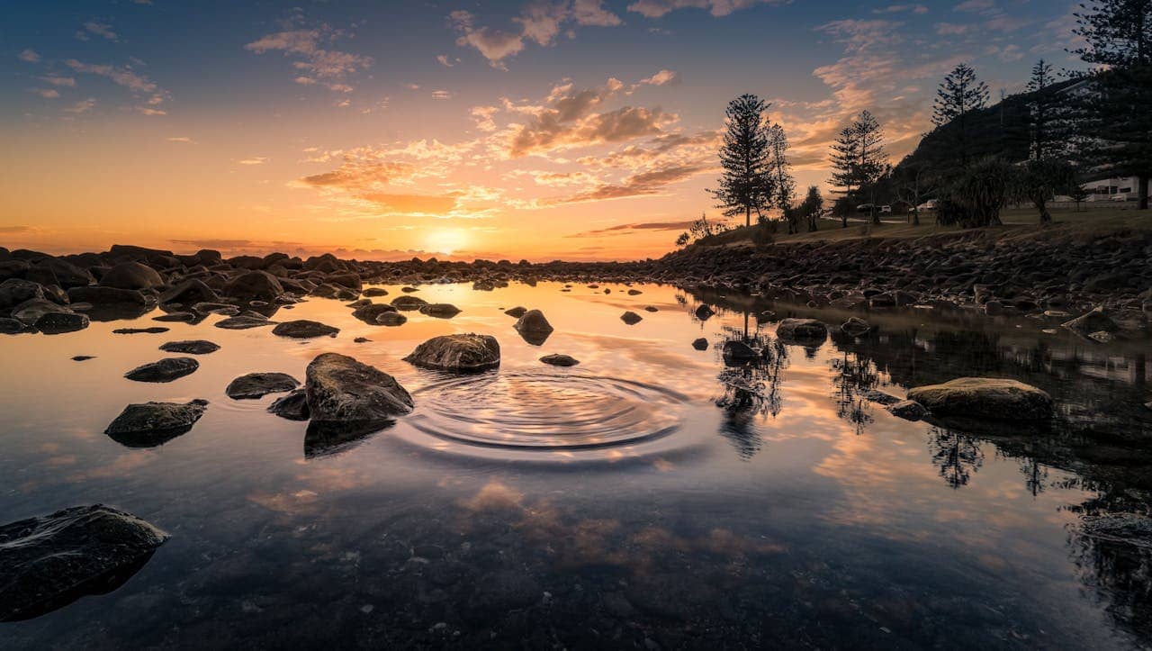 A serene sunset reflecting on a calm body of water surrounded by rocks and trees. Gentle ripples spread outward from a central point in the water, creating a peaceful and harmonious scene. The sky transitions from vibrant orange and yellow hues near the horizon to soft blues higher up, with scattered clouds adding texture.