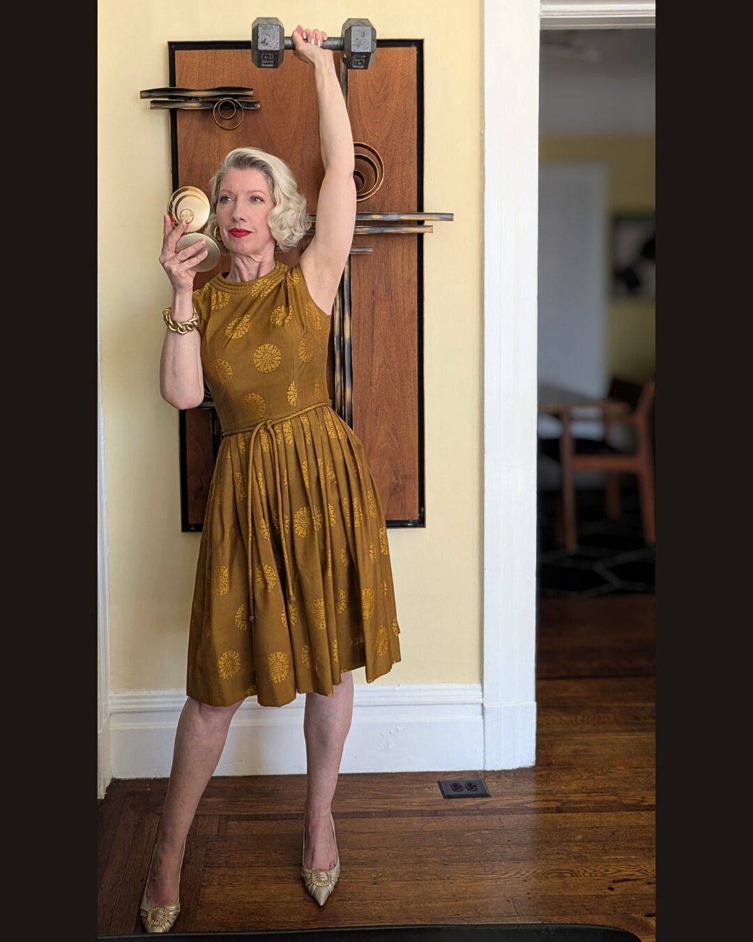 woman in gold cocktail dress holding weight overhead while checking face in compact