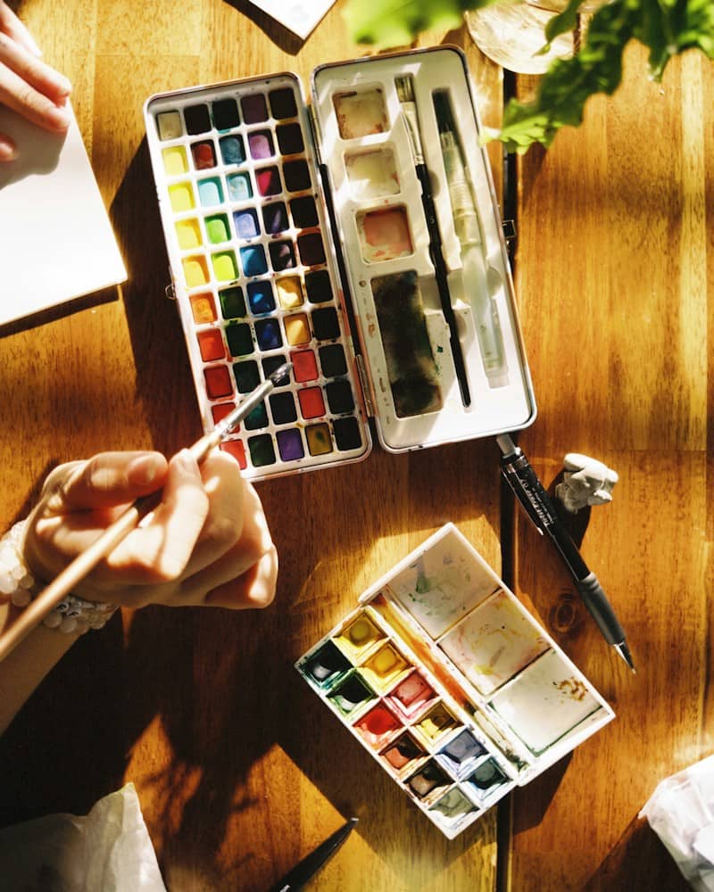 A person holding a paintbrush over watercolor pans on a sunlit wooden table