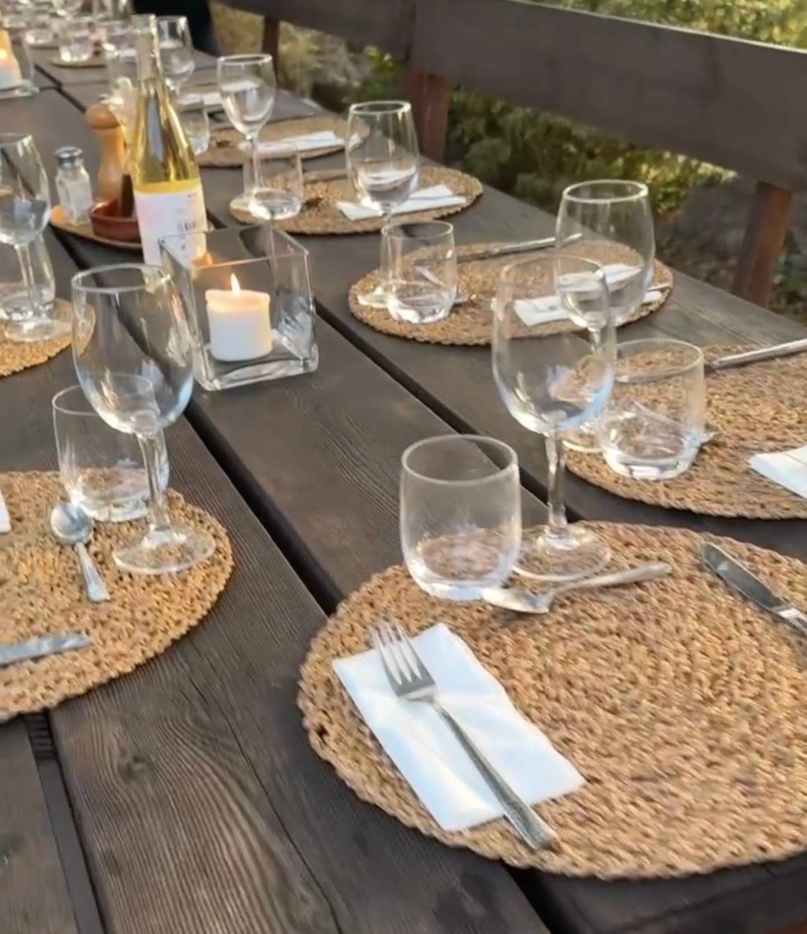 A rustic wooden table set with woven placemats, wine glasses, and a lit candle, ready for a shared meal.