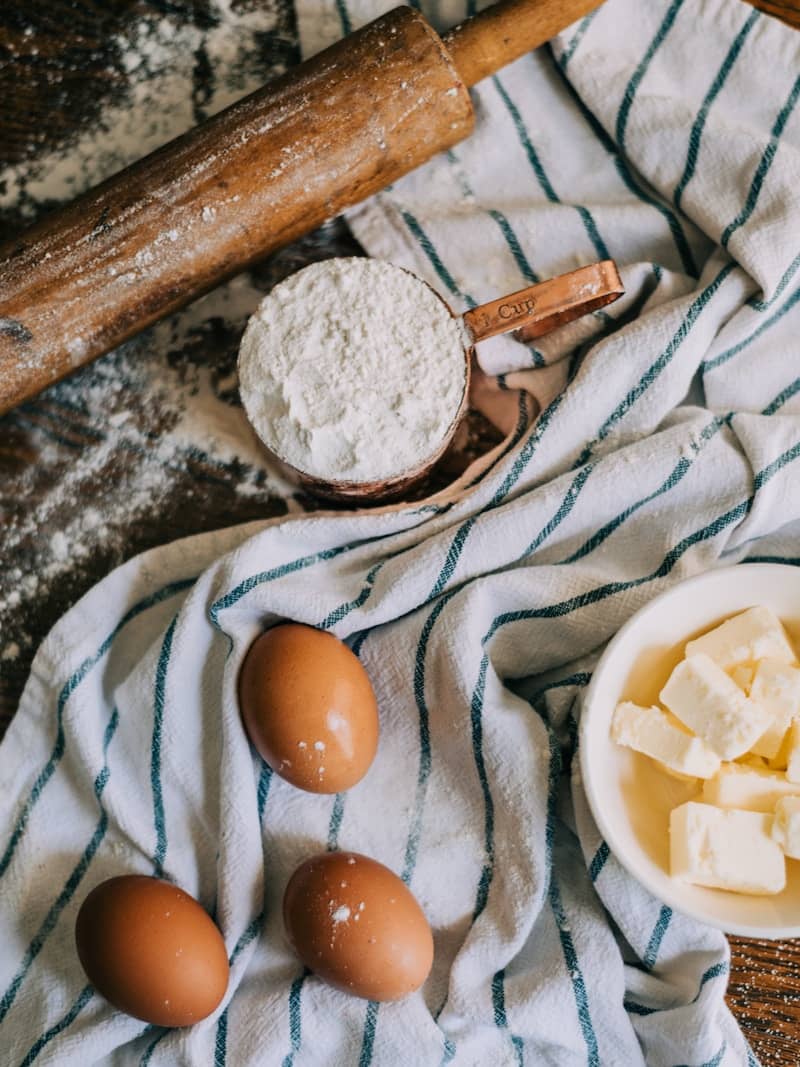 A rolling pin, flour, butter and eggs next to a tea towel.