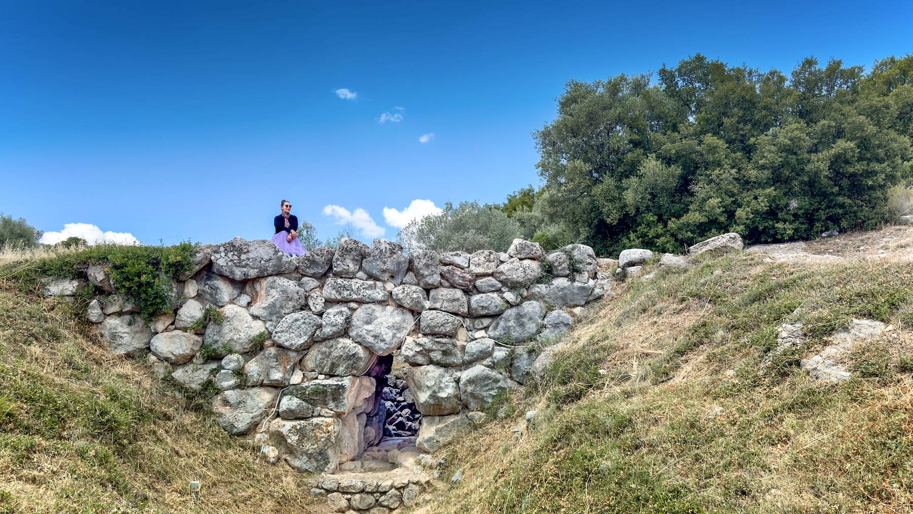Traveler sitting on top of an ancient Mycenaean stone bridge in Greece, surrounded by greenery and clear blue sky, showcasing archaeological history and cultural heritage.