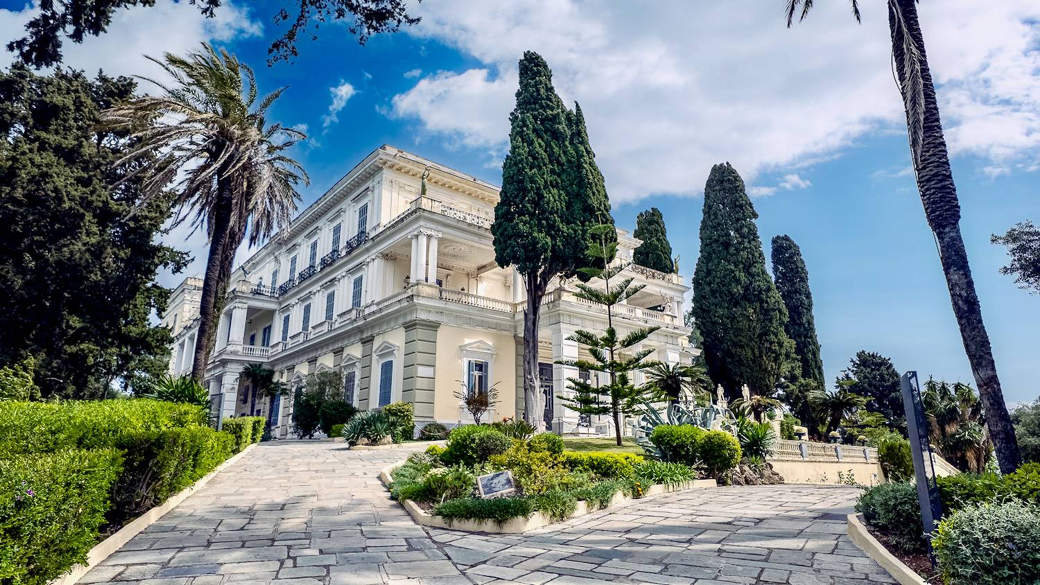 A grand neoclassical mansion with white columns and balconies surrounded by lush gardens and tall cypress and palm trees under a partly cloudy blue sky.
