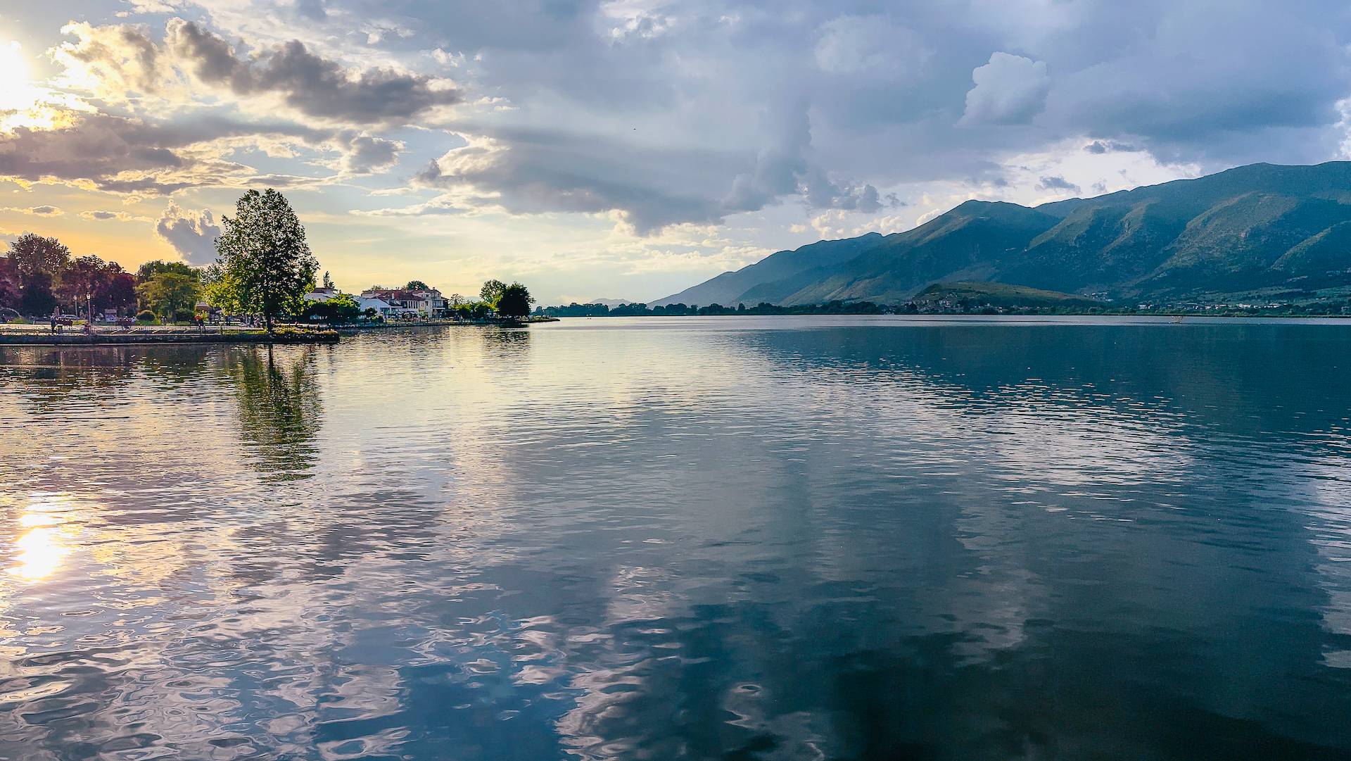 A wide view of Lake Pamvotis in Ioannina, Greece, under a dramatic sky with soft sunlight breaking through clouds at sunset. The calm water mirrors the sky, with the historic city waterfront visible on the left and lush, rolling mountains stretching acros