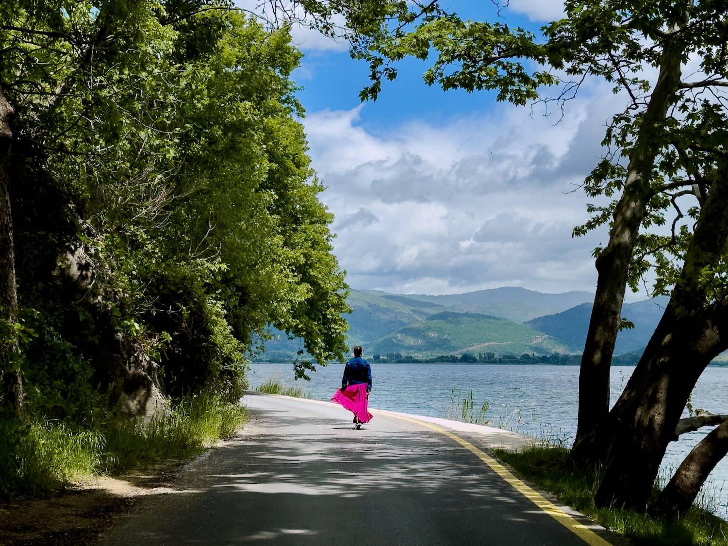 A person wearing a bright pink skirt and dark jacket walks away along a narrow lakeside road, framed by leafy trees on both sides, with calm water to the right and green mountains under a partly cloudy blue sky in the distance.
